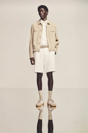 A full shot of a dark-skinned male model in light casual clothing, including a jacket, shorts, button-down shirt, socks, and shoes, posing in a studio.