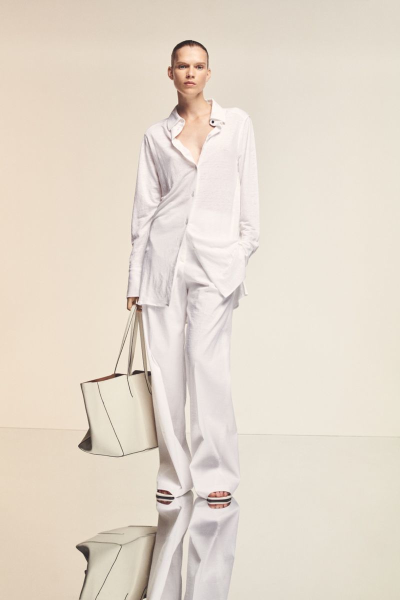 Adult woman poses in a white button-down shirt and matching white wide-leg pants, holding a tote bag against a pale gradient background.