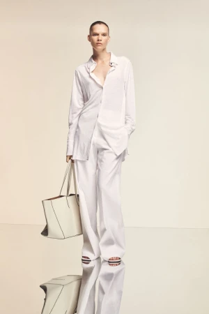 Model wearing a loose white button-down shirt and wide-leg trousers while holding a structured white tote bag on a reflective studio floor.