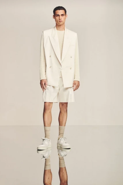 Full shot of a male model wearing a beige blazer, matching shorts, long-sleeve shirt, socks, and sneakers, posing in a photo studio.