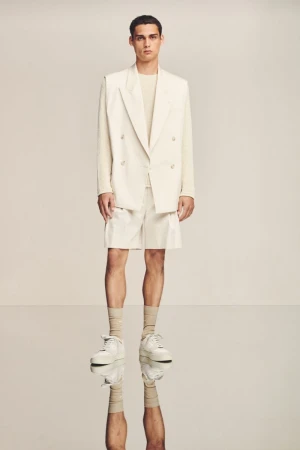 Full shot of a male model wearing a beige blazer, matching shorts, long-sleeve shirt, socks, and sneakers, posing in a photo studio.