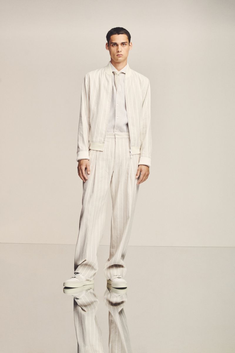 A stylish man in a cream-colored pinstripe suit poses in a studio against a neutral background, showcasing modern fashion.