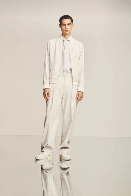 A stylish man in a cream-colored pinstripe suit poses in a studio against a neutral background, showcasing modern fashion.