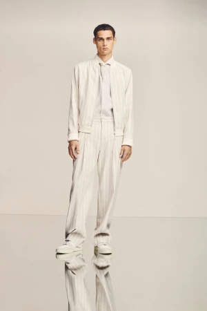 A stylish man in a cream-colored pinstripe suit poses in a studio against a neutral background, showcasing modern fashion.
