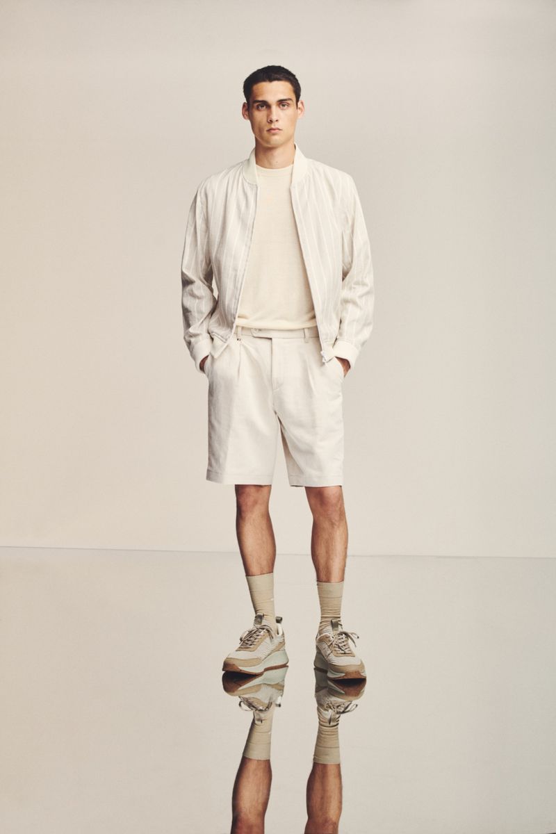 A man poses in a studio wearing a casual outfit of a bomber jacket, t-shirt, and shorts with sneakers, his reflection visible below.