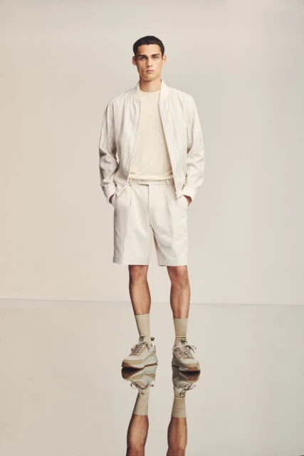 A man poses in a studio wearing a casual outfit of a bomber jacket, t-shirt, and shorts with sneakers, his reflection visible below.