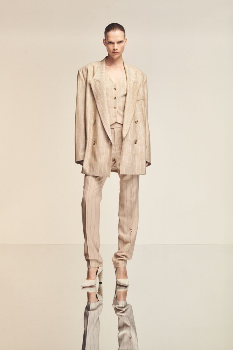 Fashionable adult female posing in a striped three-piece suit with matching heels on a reflective surface in a studio setting.