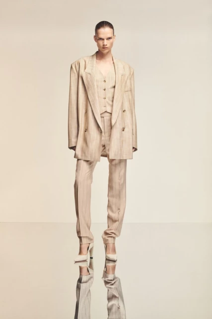 Fashionable adult female posing in a striped three-piece suit with matching heels on a reflective surface in a studio setting.