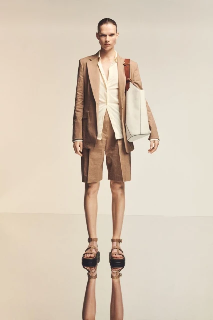 Person in beige linen suit with tote bag posing in studio with reflective floor. Fashion-forward attire and neutral color palette.