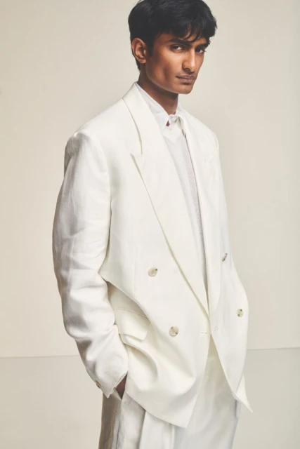 Portrait of a young adult man in a double-breasted white suit, set against a neutral backdrop, showcasing a modern, elegant fashion style.