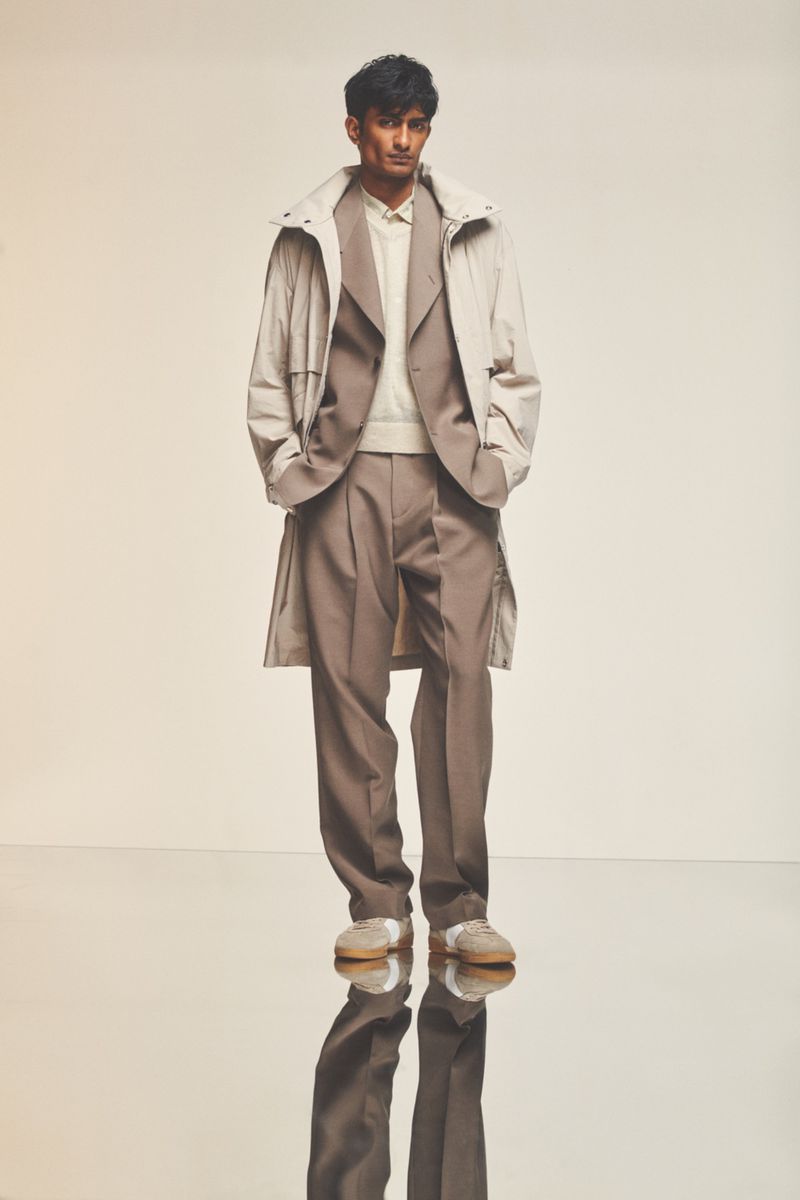 Man in a light beige overcoat and brown suit stands on reflective floor. Full-length fashion shot in front of a plain, light background.