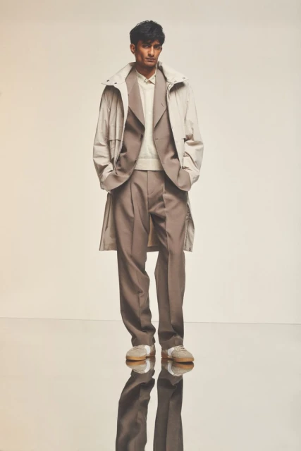 Man in a light beige overcoat and brown suit stands on reflective floor. Full-length fashion shot in front of a plain, light background.