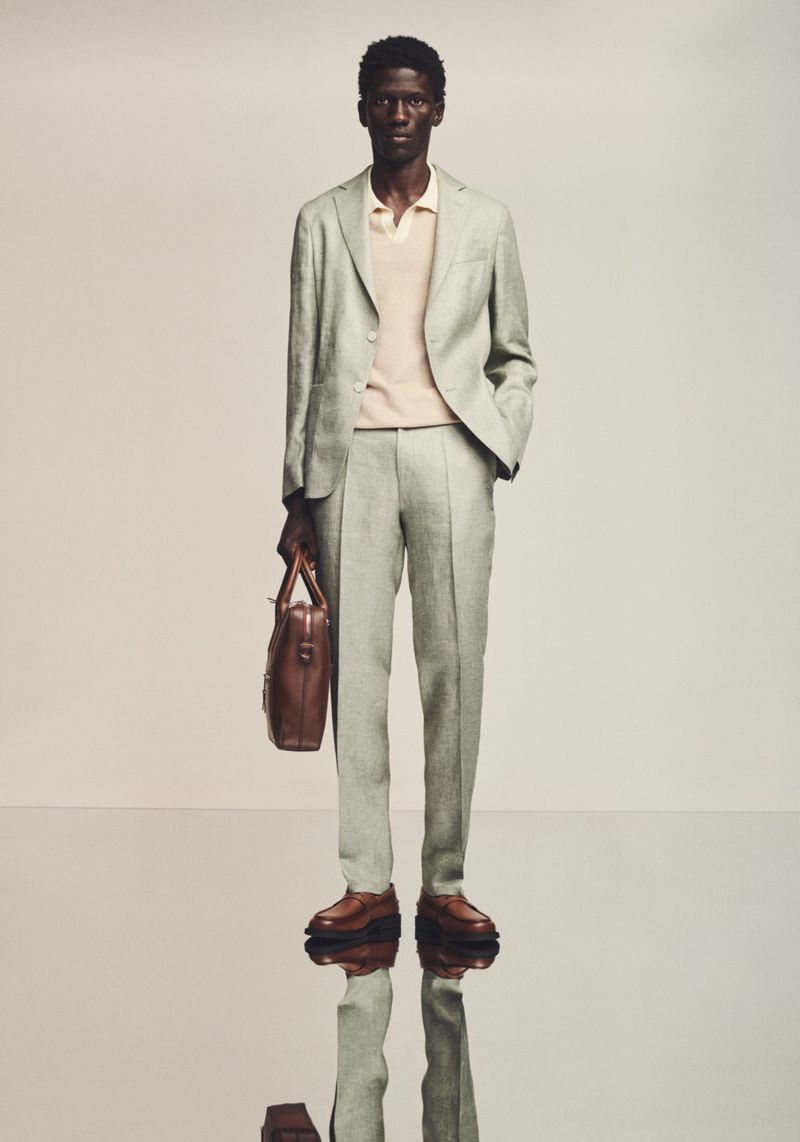 A full-length studio shot of a Black male model in a light green suit, tan polo, and brown loafers, holding a brown leather briefcase.