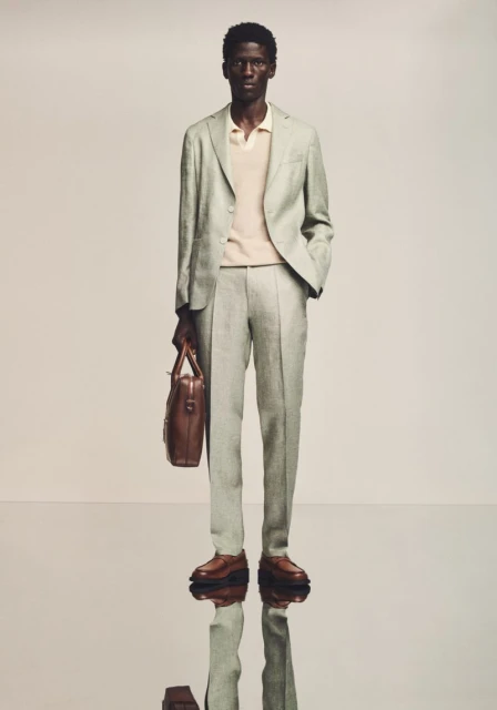 A full-length studio shot of a Black male model in a light green suit, tan polo, and brown loafers, holding a brown leather briefcase.