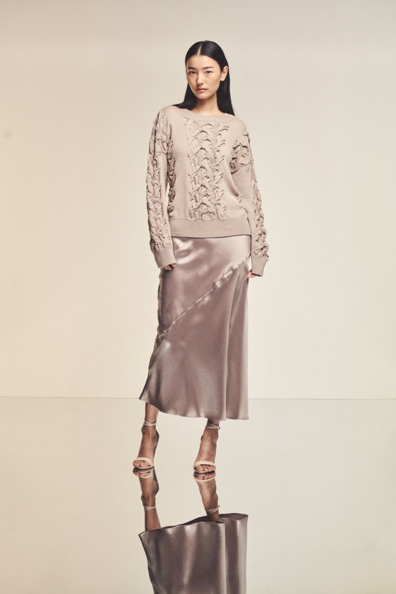 Asian woman poses in a beige knit sweater and satin skirt with high heels, reflected on the glossy floor in a neutral studio.