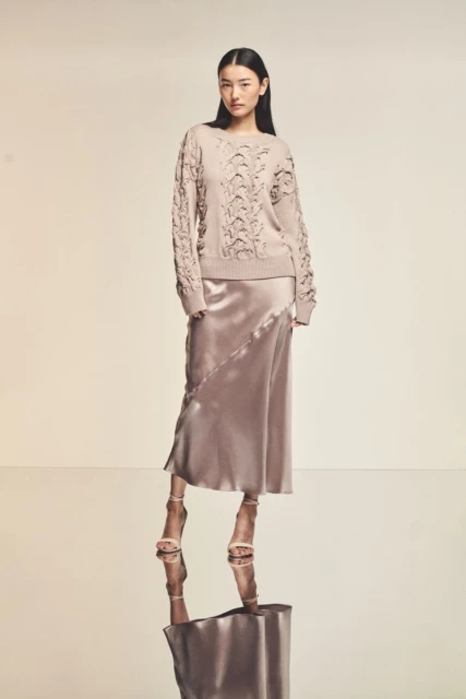 Asian woman poses in a beige knit sweater and satin skirt with high heels, reflected on the glossy floor in a neutral studio.