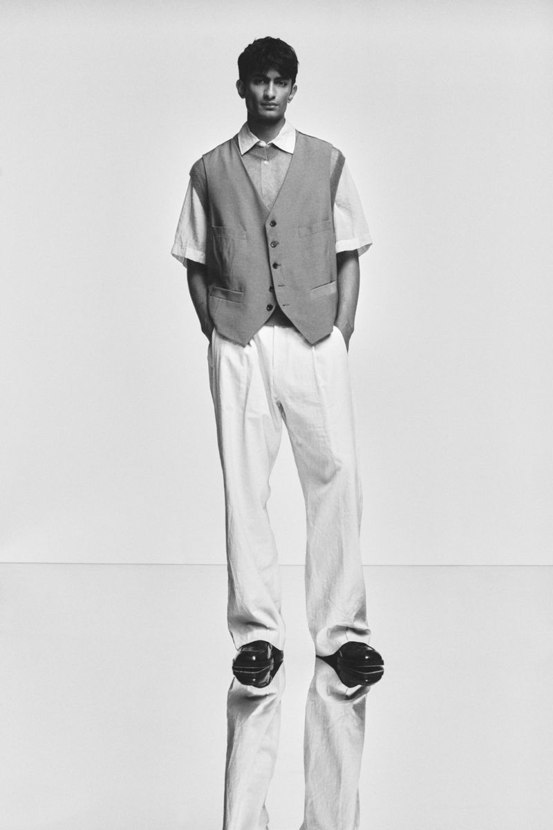 Monochrome portrait of a stylish man in a vest and pleated pants standing on a reflective floor, captured in a studio setting.