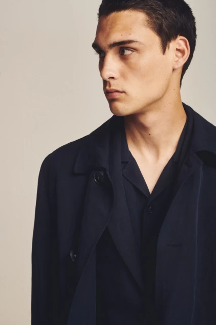 Headshot of a fair-skinned adult male model in a dark navy trench coat and collared shirt, looking off to the left side with neutral expression.