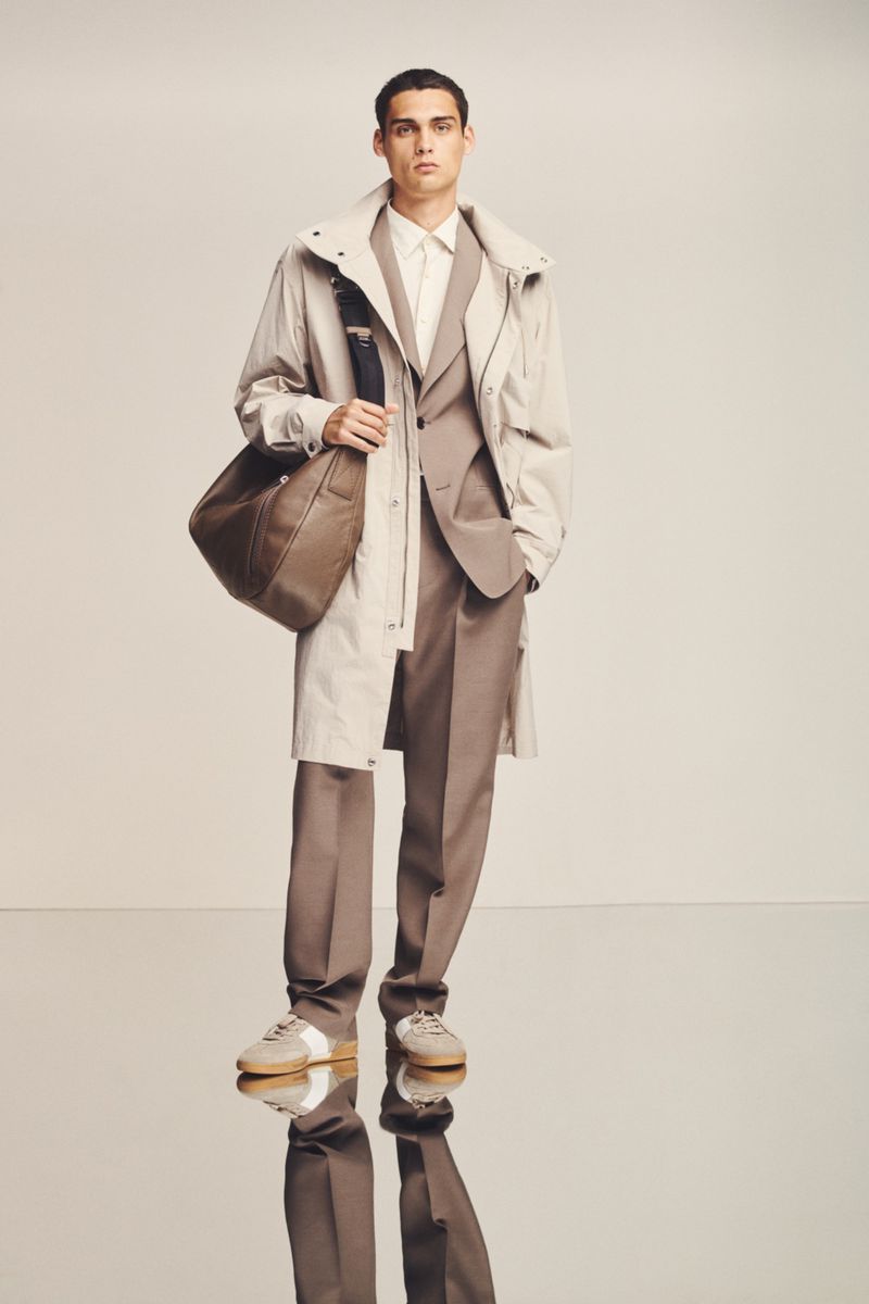 A man models a tan suit with a beige trench coat and a leather messenger bag, standing in a studio with reflective flooring.