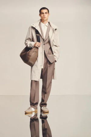 A man models a tan suit with a beige trench coat and a leather messenger bag, standing in a studio with reflective flooring.
