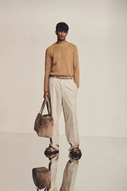 A full-length studio shot showcases a dark-haired man wearing a tan sweater, cream slacks, and sandals, carrying a tote bag.