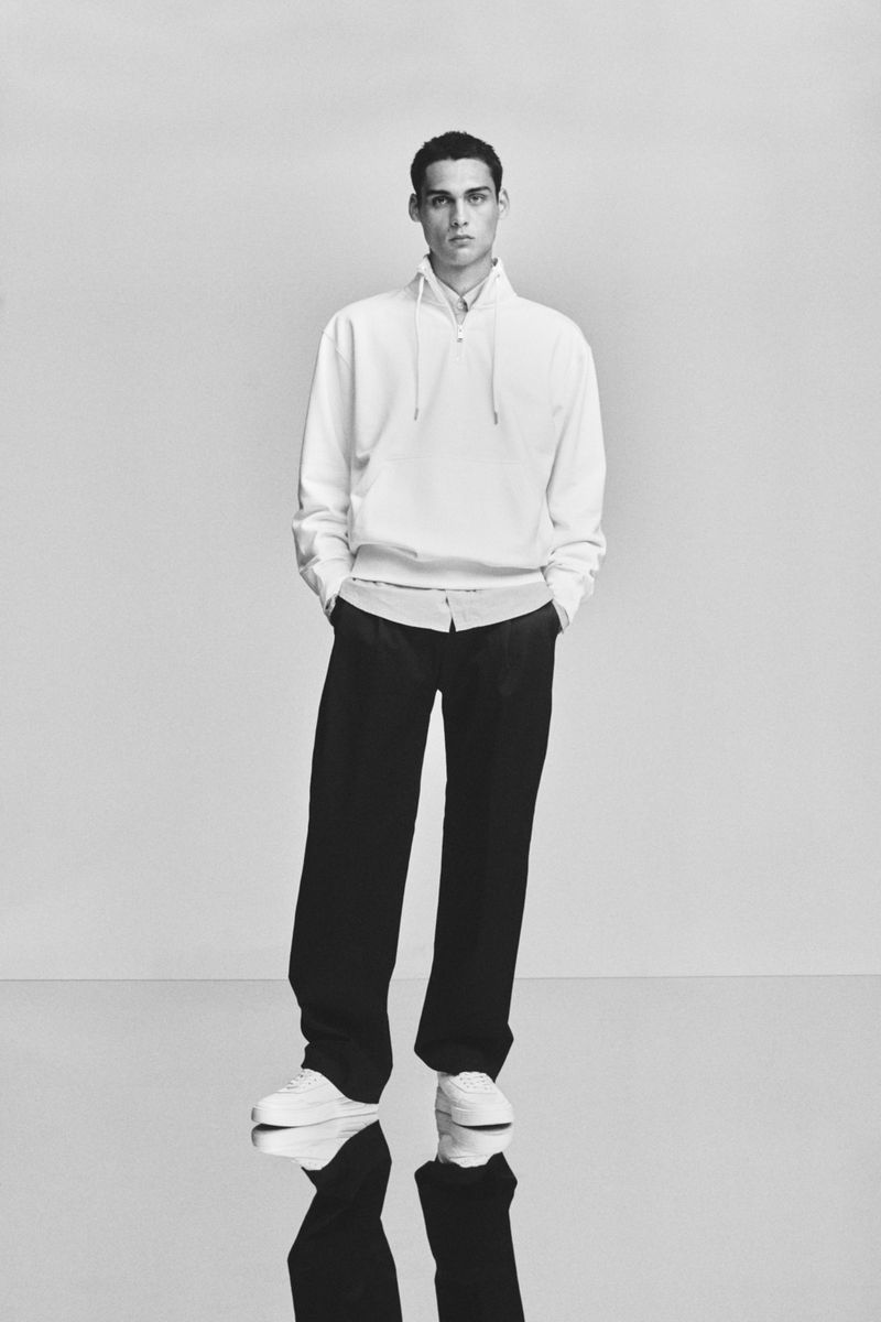 Monochrome full shot of a young adult male model standing in a white hooded sweatshirt, dress shirt, dress pants, and sneakers.