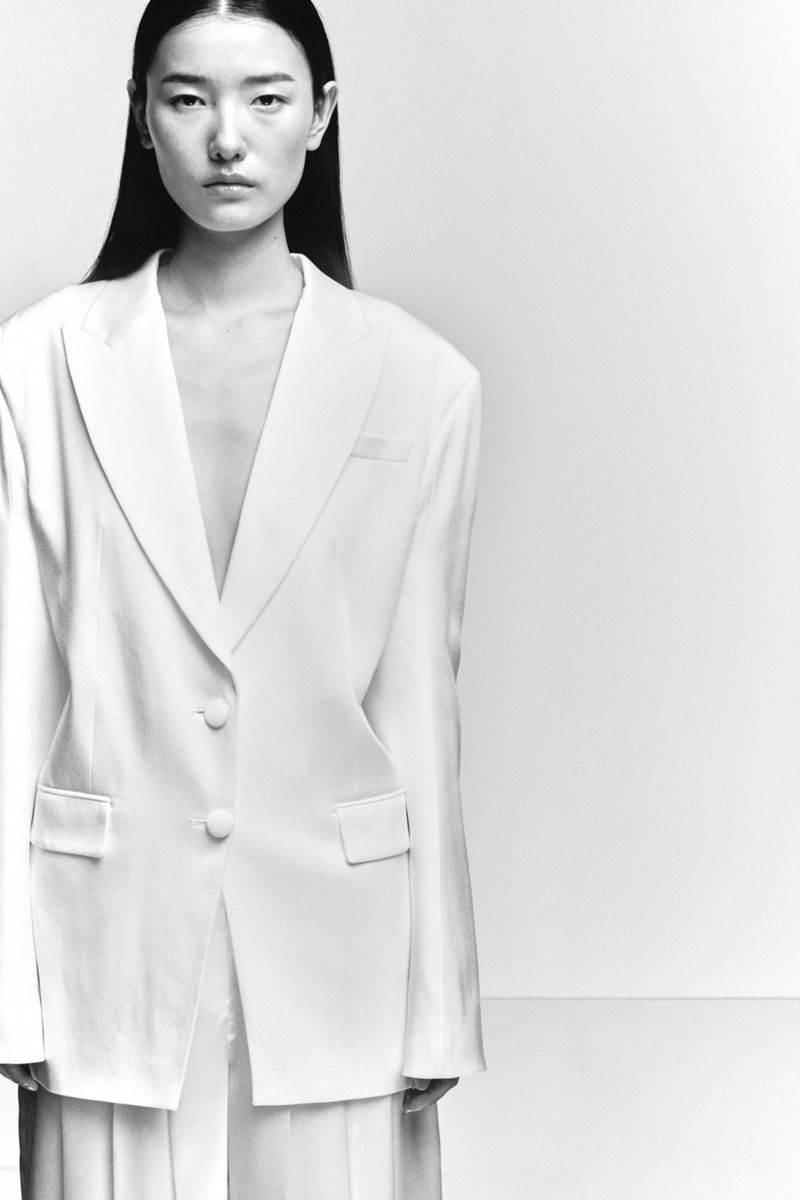Asian woman in an oversized white blazer with straight black hair, shot in a white studio with bright lighting and a neutral expression.