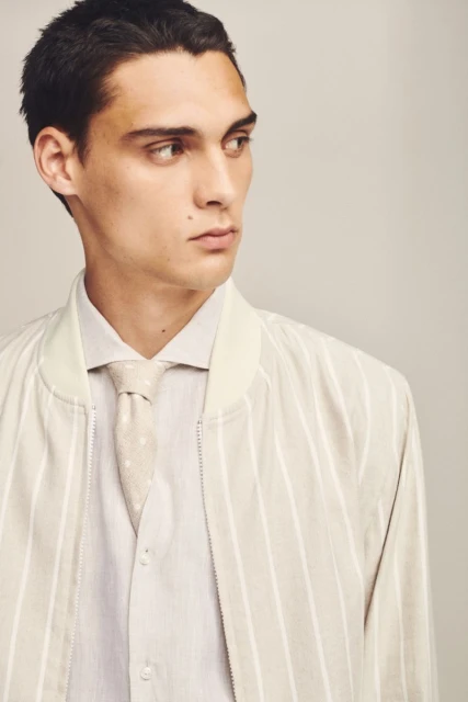 Portrait of a young adult male in a striped jacket, light shirt, and patterned tie, looking off-camera in a studio setting.