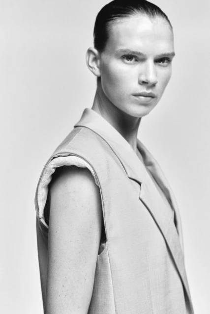 Black and white portrait of androgynous adult with short hair, wearing a tailored vest against a neutral background, looking at the camera.