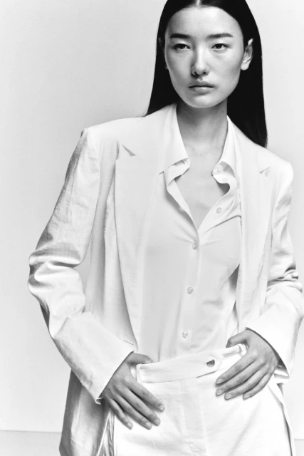 Black-and-white portrait of a woman in a white linen blazer and button-down shirt, hands resting on her waist. Fashion editorial.
