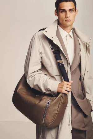 Fashion shot of a male model in a beige trench coat and tan suit, carrying a brown leather messenger bag in a studio setting.