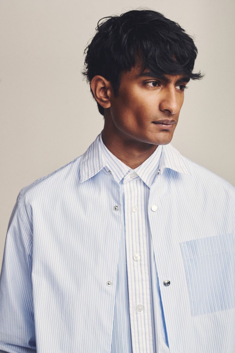 Headshot of a dark-haired male model wearing a button-down striped shirt and an unbuttoned light-blue striped overshirt indoors.