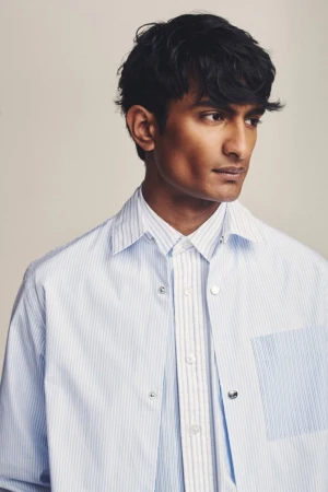 Headshot of a dark-haired male model wearing a button-down striped shirt and an unbuttoned light-blue striped overshirt indoors.