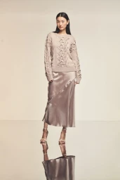 Model wearing a beige patterned knit sweater and satin midi skirt on a glossy reflective floor against a neutral background.