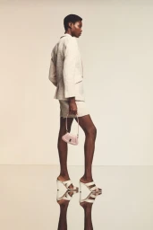 Model in a cream tailored blazer and shorts holding a small pink handbag standing in profile on a reflective studio floor.