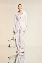 Model wearing a loose white button-down shirt and wide-leg trousers while holding a structured white tote bag on a reflective studio floor.