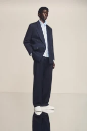 Black male model wearing an oversized navy double-breasted suit, light blue shirt, and white sneakers against a neutral backdrop.