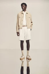 Model standing on a reflective studio floor wearing a beige jacket, white shirt, white shorts, socks, and sneakers.