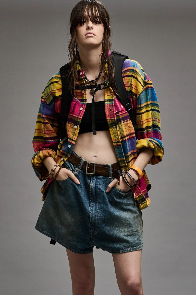 A young woman with wet hair, wearing a plaid shirt and denim shorts, poses in a studio. She carries a black backpack.