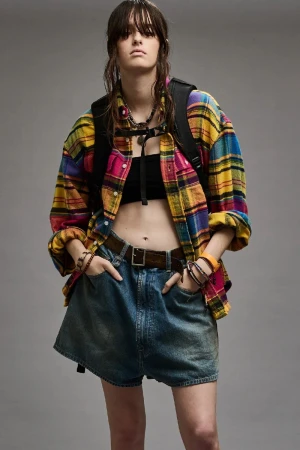 A young woman with wet hair, wearing a plaid shirt and denim shorts, poses in a studio. She carries a black backpack.