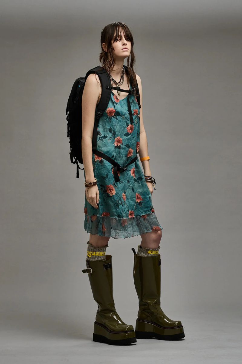 A young woman stands in a studio wearing a floral dress and platform boots, carrying a black backpack against a neutral backdrop.