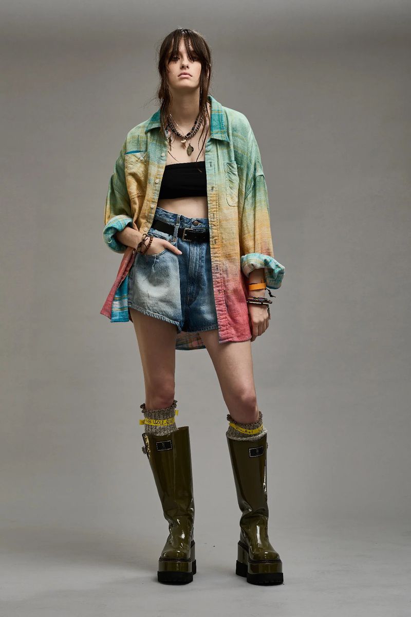 Adult woman wearing colorful shirt, denim shorts, and high boots in a studio fashion shot with a neutral background, looking at the camera.