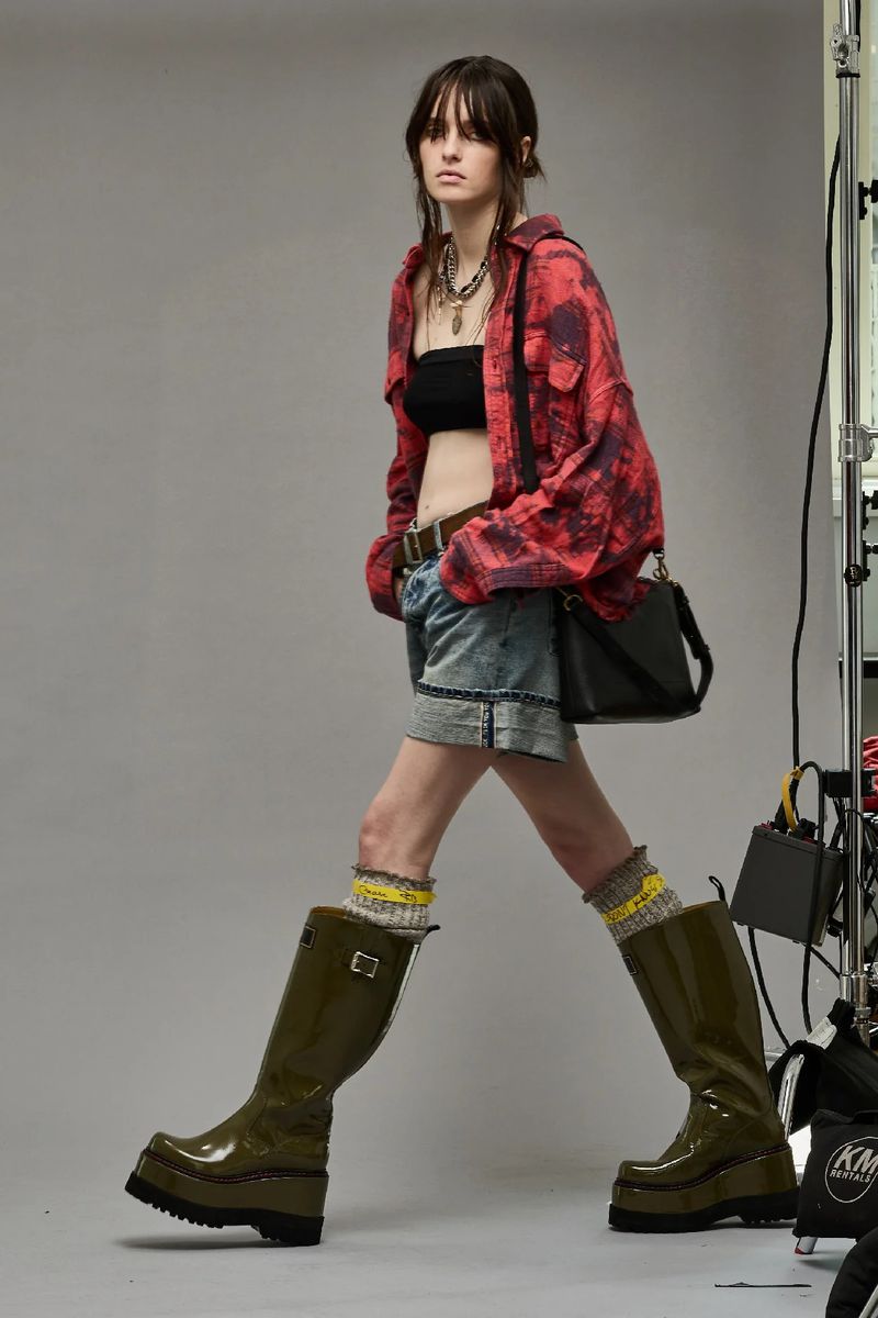 A young woman with bangs and dark hair stands confidently in a studio, wearing a black tube top, denim shorts, and glossy olive green boots.