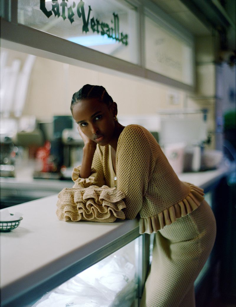 Woman in a beige knitted outfit leans on a counter in front of a cafe, with a ruffled handbag, bathed in soft, natural light.