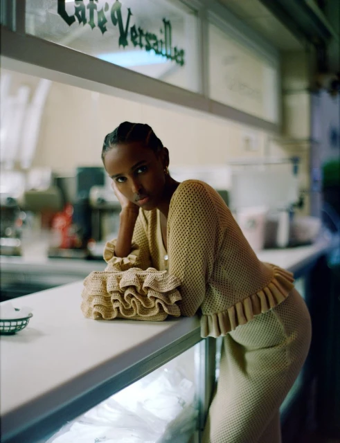 Woman in a beige knitted outfit leans on a counter in front of a cafe, with a ruffled handbag, bathed in soft, natural light.
