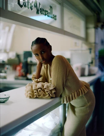Woman in a beige knitted outfit leans on a counter in front of a cafe, with a ruffled handbag, bathed in soft, natural light.