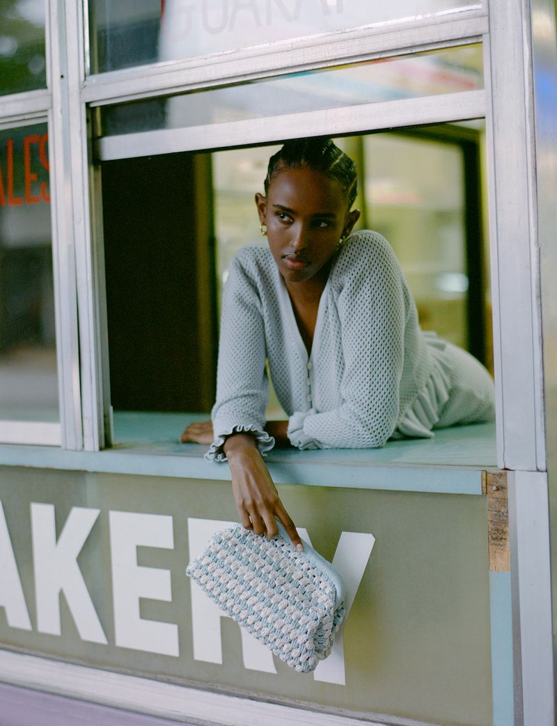 Portrait of a stylish young adult female leaning out of a bakery window, holding a blue woven handbag, wearing a blue cardigan.