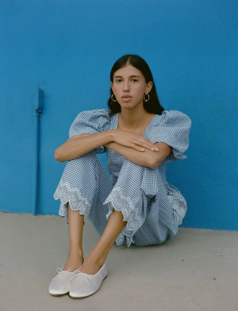 A young woman sits with arms crossed, wearing a blue and white gingham top and pants set with white ballet flats against a solid blue wall.