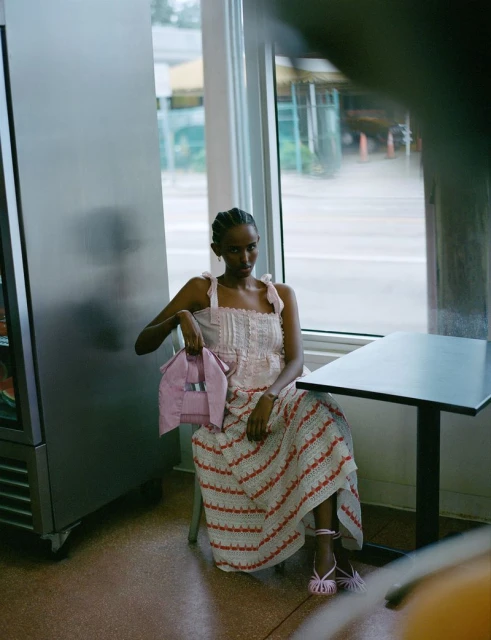 A woman in a patterned dress sits in front of a window with a pink jacket draped over her bag, illuminated by natural light.