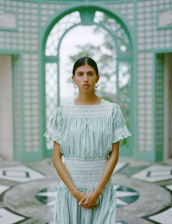 Portrait of a young woman with green-and-white striped dress in front of a light green garden pavilion with decorative tile flooring.